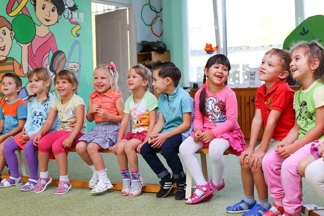 Kisgyerekek ülnek egy padon egy óvodai csoportszobában, mosolyogva néznek jobbra, mögöttük színes falfestés, ajtó és nagy ablakok.