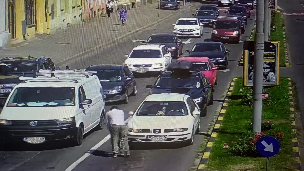 Térfigyelő kamera felvétele forgalmas városi utcáról, ahol több autó áll sorban egymás mögött. Az úttest közepén egy férfi guggol egy fehér autó mellett, mögötte egy fehér furgon és egy kisebb autó áll.