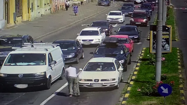 Térfigyelő kamera felvétele forgalmas városi utcáról, ahol több autó áll sorban egymás mögött. Az úttest közepén egy férfi guggol egy fehér autó mellett, mögötte egy fehér furgon és egy kisebb autó áll.