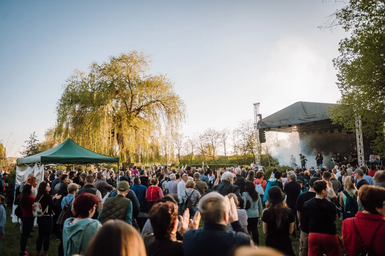 Nagy tömeg gyűlik össze egy szabadtéri színpad előtt egy parkban, a színpadon zenészek lépnek fel, mellette zöld sátor és fák láthatók, a résztvevők tapsolnak.