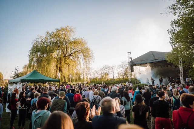 Nagy tömeg gyűlik össze egy szabadtéri színpad előtt egy parkban, a színpadon zenészek lépnek fel, mellette zöld sátor és fák láthatók, a résztvevők tapsolnak.