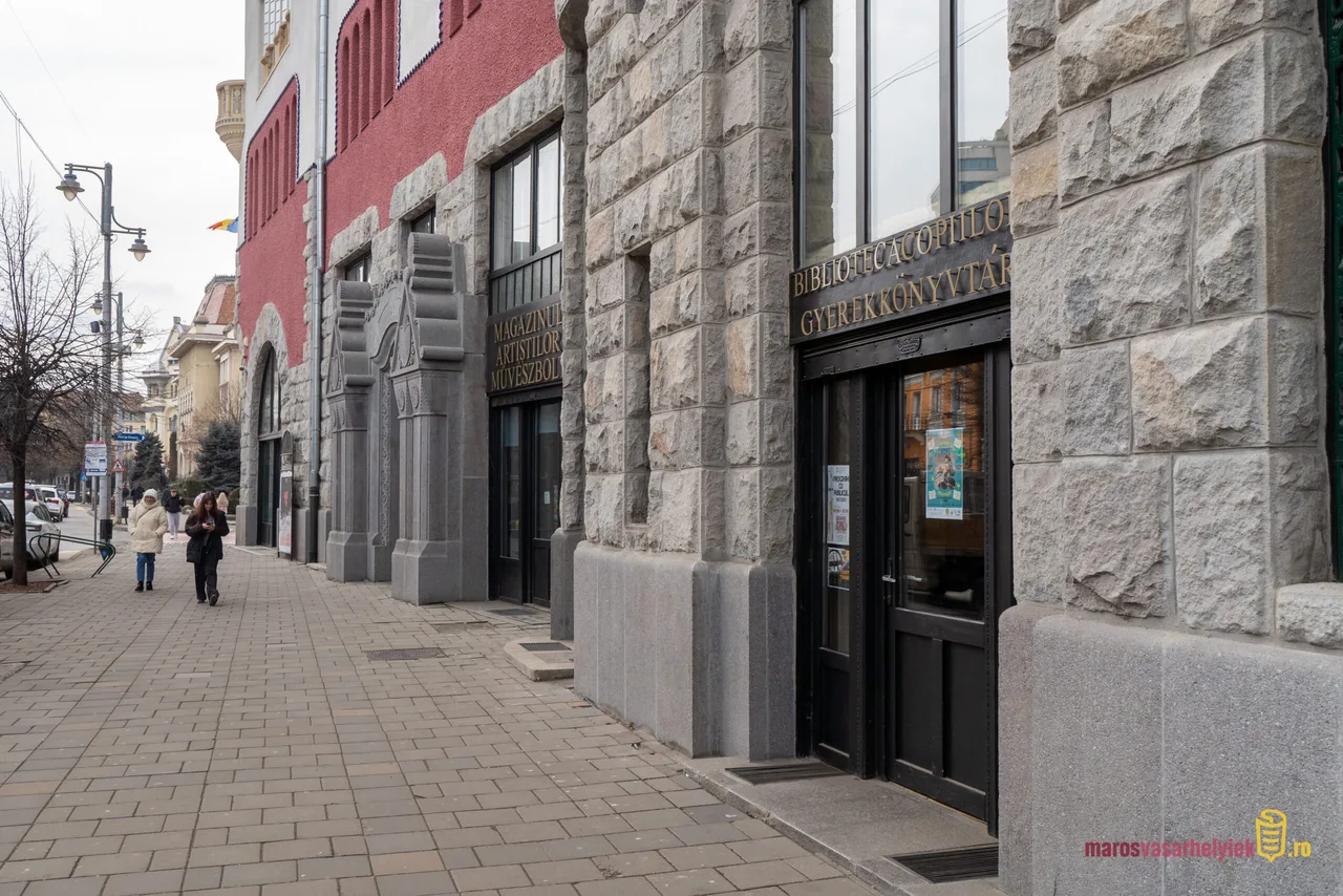 Kultúrpalota kőből épült homlokzatának részlete Marosvásárhelyen, előtte a Gyerekkönyvtár (Biblioteca Copiilor) bejárata és a Művészbolt felirat, a járdán két gyalogos sétál.