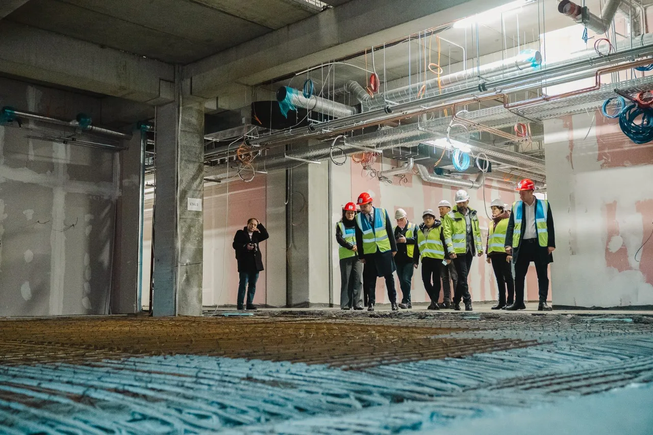 Egy csoport sárga láthatósági mellényt és sisakot viselő ember sétál egy épülő épület nyers betonszerkezetű belső terében, előtérben vasalt betonalap, felül látszó gépészeti csővezetékek és kábelek.
