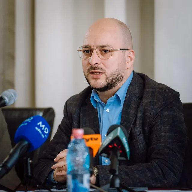 Kopasz, szemüveges, szakállas férfi sajtótájékoztatón beszél, előtte az asztalon több riportermikrofon és egy ásványvizes palack látható.