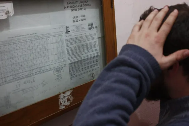 Egy férfi hátulról fotózva, kezét a fejéhez emelve néz egy üvegezett hirdetőtáblát, amelyen kimutatások és egy románul íródott hirdetmény (nyugalmi időszakra vonatkozó szabályok) láthatók.