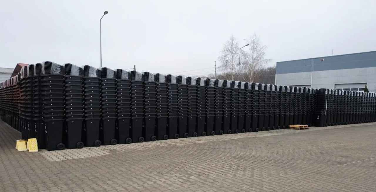 Nagy udvaron hosszú sorokban, egymásba rakva több száz fekete kerekes szemetes kuka várakozik kiosztásra, a háttérben ipari épület és lombtalan fák láthatók.
