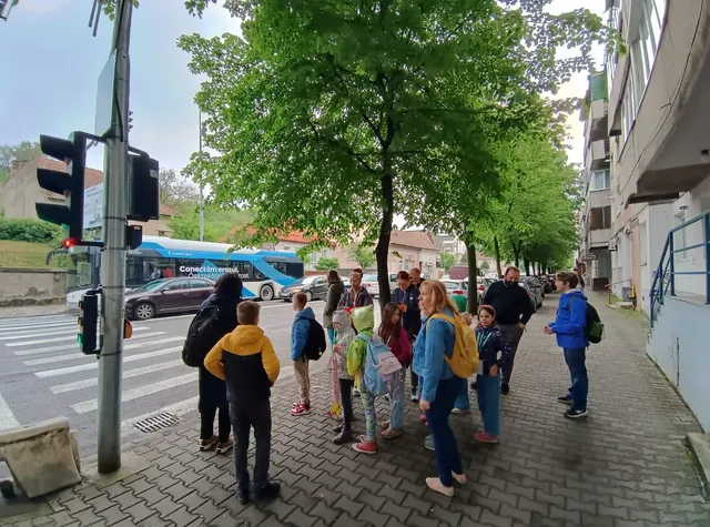 Gyerekek csoportja hátizsákkal, kísérő felnőttekkel várakozik egy fás járdaszakaszon a gyalogátkelő közelében, a háttérben elektromos busz és forgalom látható.
