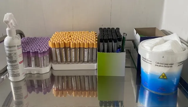 Fényes fémasztalon lila és sárga kupakos vérvételi kémcsövek sorakoznak tartókban, mellettük tollak egy zöld tartóban, egy fertőtlenítő spray, egy karton doboz és egy kék-fehér veszélyeshulladék-gyűjtő tartály vattával.