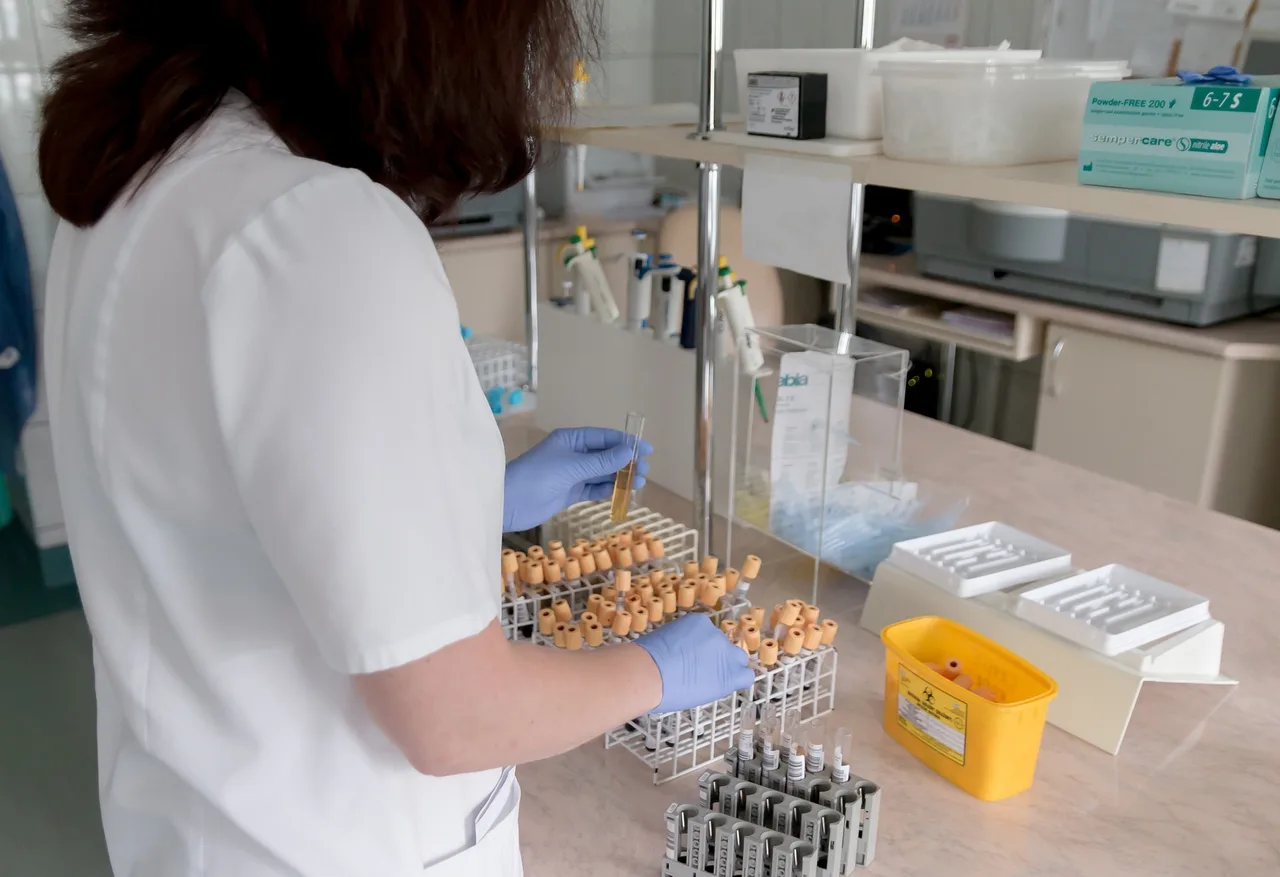 Fehér köpenyt és kék gumikesztyűt viselő laboráns vérmintás csöveket helyez el egy állványban egy laboratóriumi pult mellett, körülötte pipetták, tálcák és veszélyeshulladék-gyűjtő doboz látható.