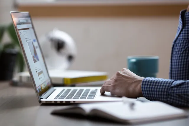 Egy férfi kockás inget visel, és íróasztalnál ülve laptop billentyűzetén, illetve érintőpadján dolgozik. Az asztalon nyitott jegyzetfüzet, egy csésze és háttérben egy fehér lámpa látható.