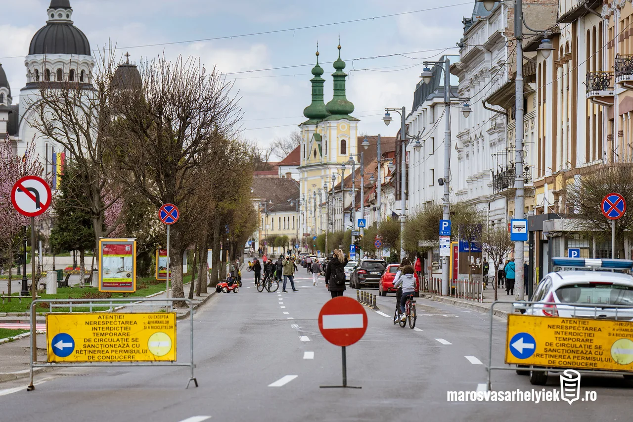 Marosvásárhely főutcájának lezárt szakasza, forgalomkorlátozó táblákkal és behajtani tilos jelzéssel, háttérben a Vártemplom tornyaival, gyalogosok és kerékpárosok sétálnak az úttesten.
