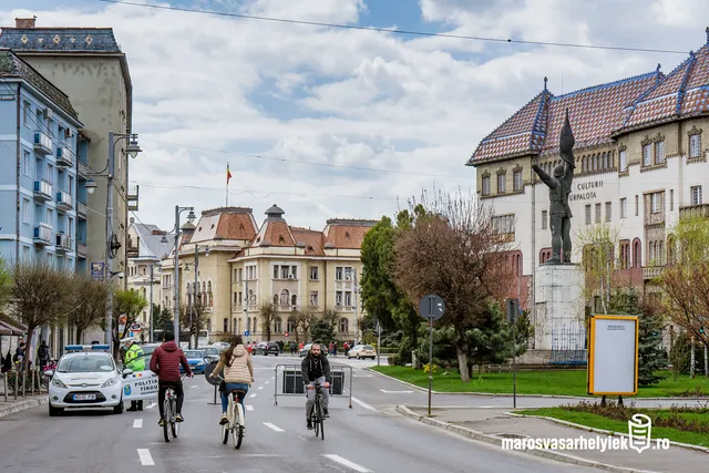 Utcarészlet Marosvásárhely belvárosában, három kerékpáros halad az úton, a háttérben a Kultúrpalota és más régi épületek láthatók, jobbra egy rendőrautó és egy rendőr áll az útlezárásnál.