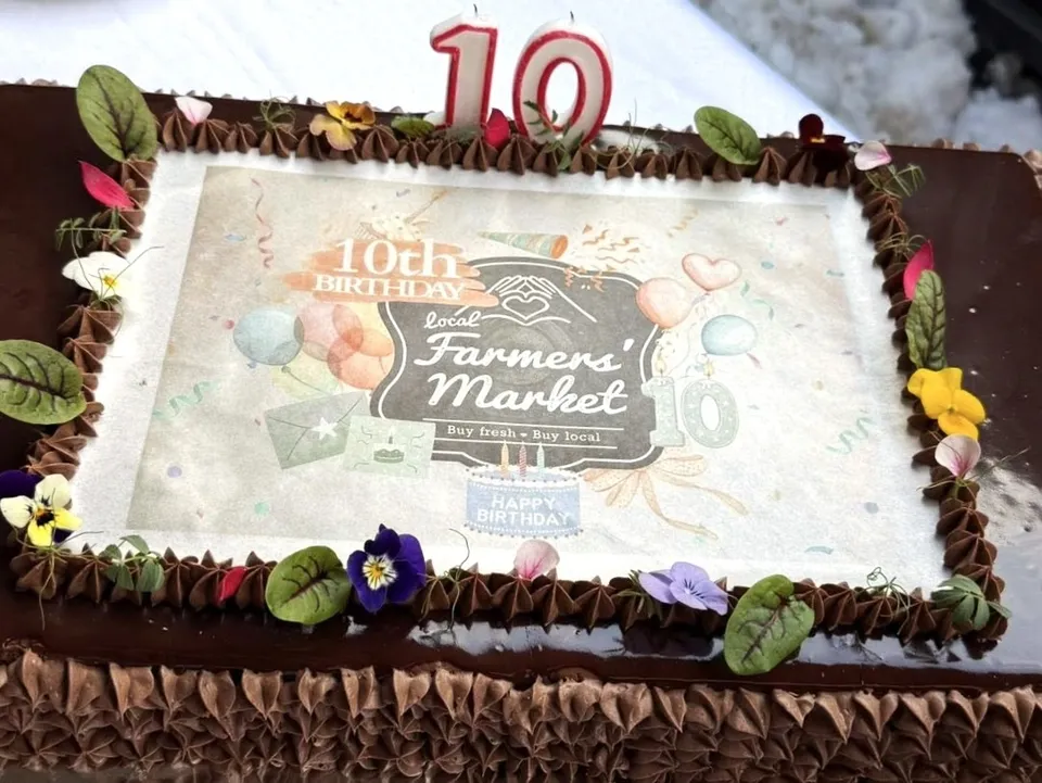 Csokoládés torta tetején ehető papírra nyomott „10th Birthday Local Farmers' Market” feliratú dekoráció, körülötte krémrózsákkal és ehető virágokkal díszítve, tetején piros 10-es alakú gyertya.