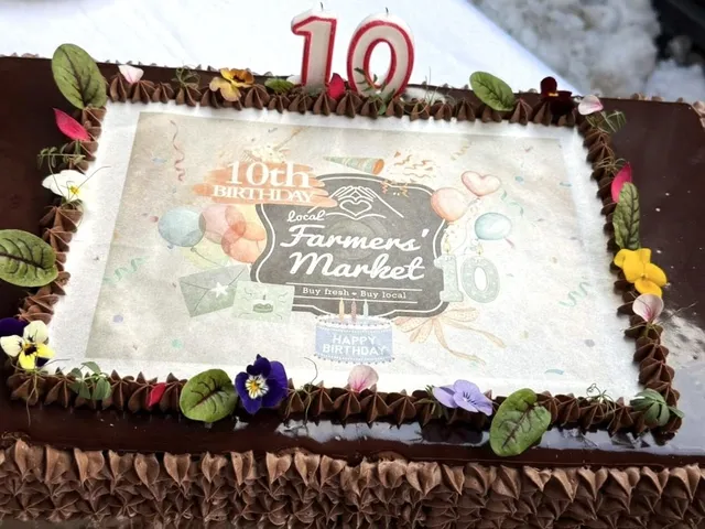 Csokoládés torta tetején ehető papírra nyomott „10th Birthday Local Farmers' Market” feliratú dekoráció, körülötte krémrózsákkal és ehető virágokkal díszítve, tetején piros 10-es alakú gyertya.