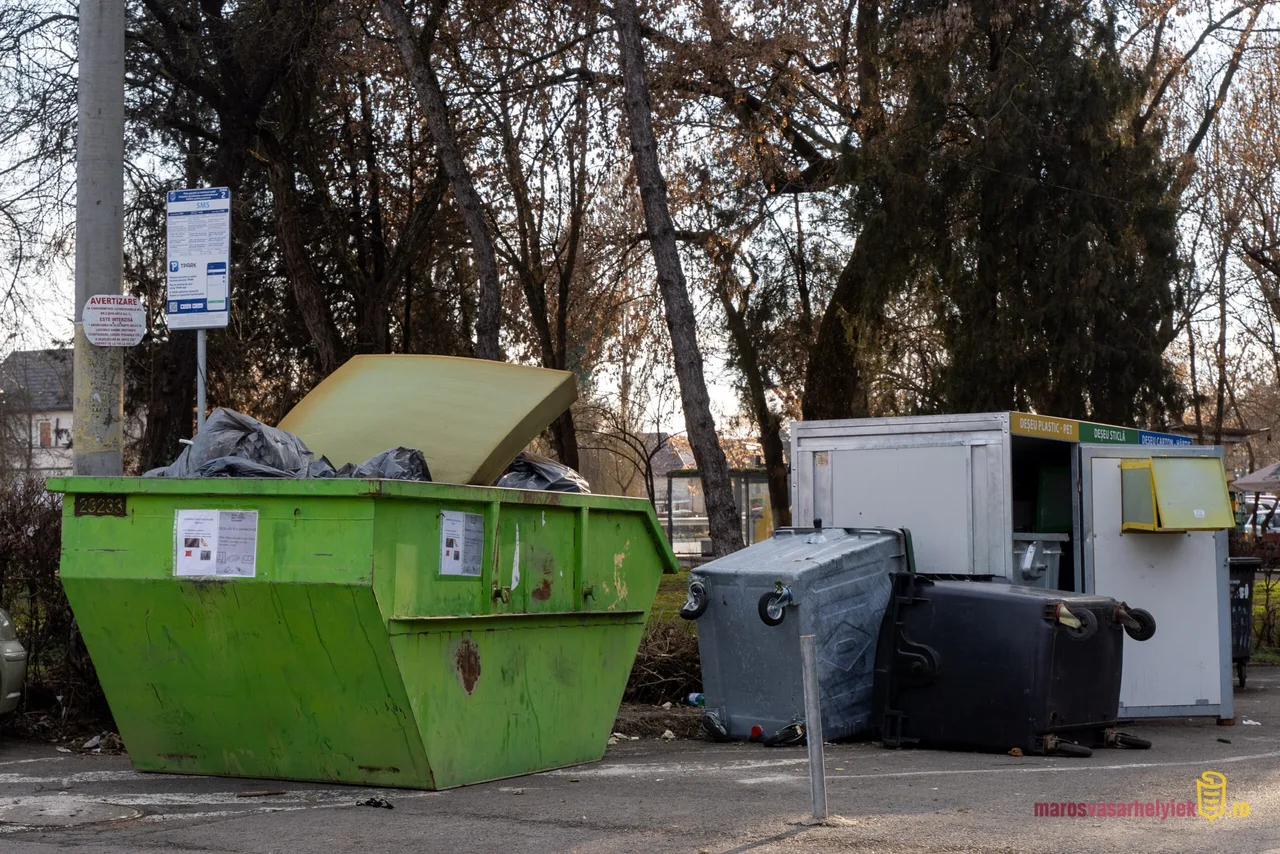 Zöld fém konténer túltelt szeméttel, mellette szelektív hulladékgyűjtő edények és egy szürke szelektív gyűjtőpont állnak egy parkolóban, háttérben fás terület látható.
