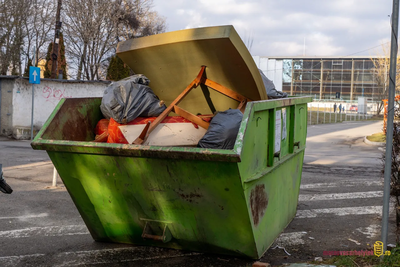 Zöld színű, nagy méretű lomtalanítási konténer utcán, tele hulladékkal: szemeteszsákok, egy fadarab és egyéb kidobott tárgyak, háttérben lakóházak és egy nagyobb üvegfalú épület látható.