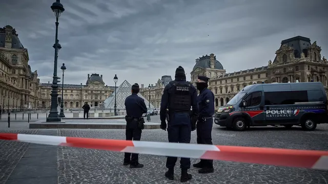 Rendőrségi elzárószalag mögött francia rendőrök állnak a párizsi Louvre múzeum udvarán, a háttérben az üvegpiramissal és a múzeum épületével, mellettük rendőrségi kisbusz parkol.