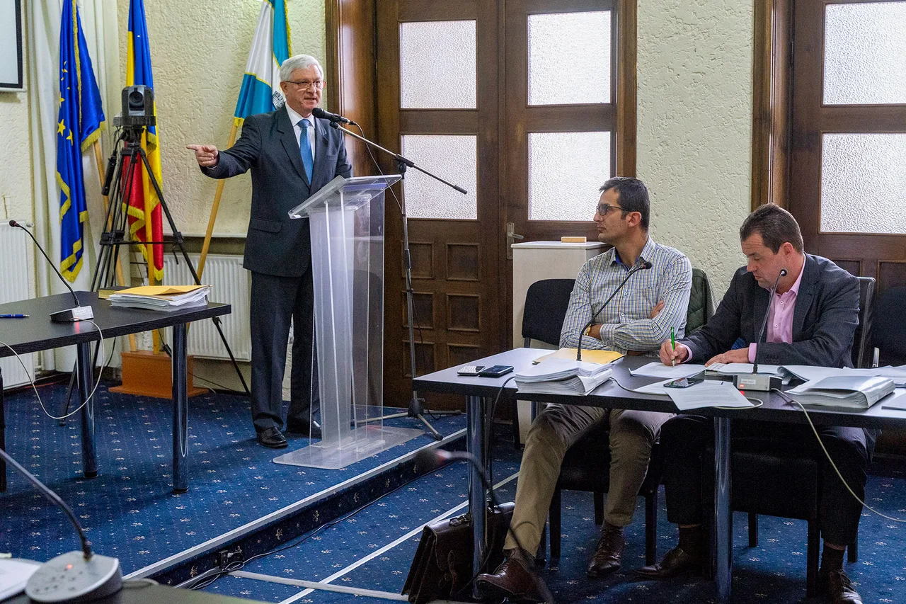 Tanácsteremben egy idősebb férfi áll a pulpitusnál, mikrofonba beszél és kézzel mutat valamerre, mögötte az EU, román és székely zászló látható. Előtérben egy asztalnál két férfi ül, dokumentumokkal és mikrofonokkal, figyelik az előadót.