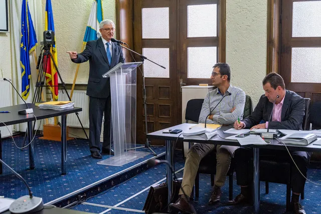 Tanácsteremben egy idősebb férfi áll a pulpitusnál, mikrofonba beszél és kézzel mutat valamerre, mögötte az EU, román és székely zászló látható. Előtérben egy asztalnál két férfi ül, dokumentumokkal és mikrofonokkal, figyelik az előadót.