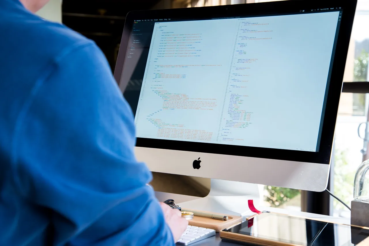 Egy kék pulóveres személy háttal ül egy iMac asztali számítógép előtt, a képernyőn programkód (HTML/CSS) látható.