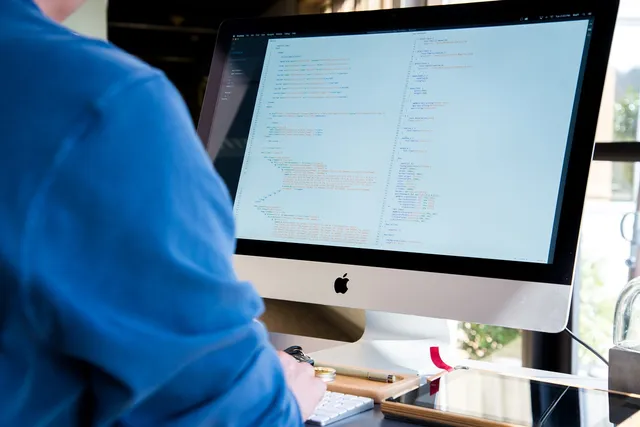 Egy kék pulóveres személy háttal ül egy iMac asztali számítógép előtt, a képernyőn programkód (HTML/CSS) látható.