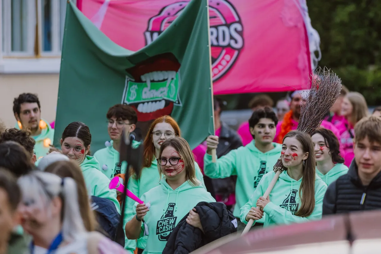 Fiatalok csoportja vonul az utcán azonos színű, mentazöld pulóverekben, sebhelyeket és vért utánzó arcfestéssel, a háttérben egy nagy zöld zászlót és rózsaszín transzparenst tartanak fel.