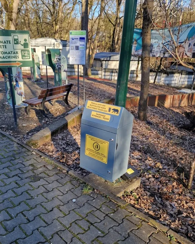 Egy medvebiztos, zárható fém szemeteskuka áll egy parkosított séta útnál, mellette pad, információs táblák és jegyautomata-jelzés láthatók, háttérben lombtalan fák és állatkerti kerítés.