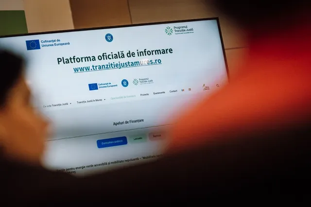 Számítógép-monitor képernyője, rajta a „Tranziție Justă Mureș” (Méltányos Átállás Program Maros megye) hivatalos tájékoztató weboldala látható, előtte homályosan egy személy keze és karja.