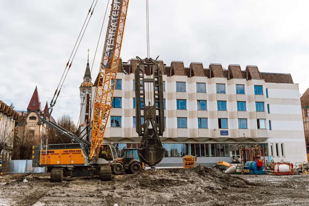 Nagy méretű sárga darukotró és markológép dolgozik egy építési területen, háttérben egy több emeletes fehér-bézs csíkozott homlokzatú épülettel, a távolban a Vártemplom vagy hasonló toronyépület látható.