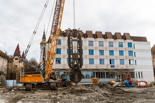 Nagy méretű sárga darukotró és markológép dolgozik egy építési területen, háttérben egy több emeletes fehér-bézs csíkozott homlokzatú épülettel, a távolban a Vártemplom vagy hasonló toronyépület látható.