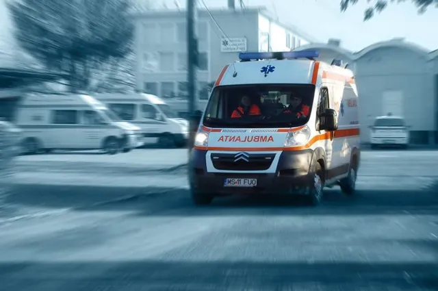 Mozgásélességgel fotózott mentőautó halad úton, elöl AMBULANTA felirattal és villogó jelzőfényekkel, a vezetőfülkében két, narancssárga mentős egyenruhát viselő személy látható.