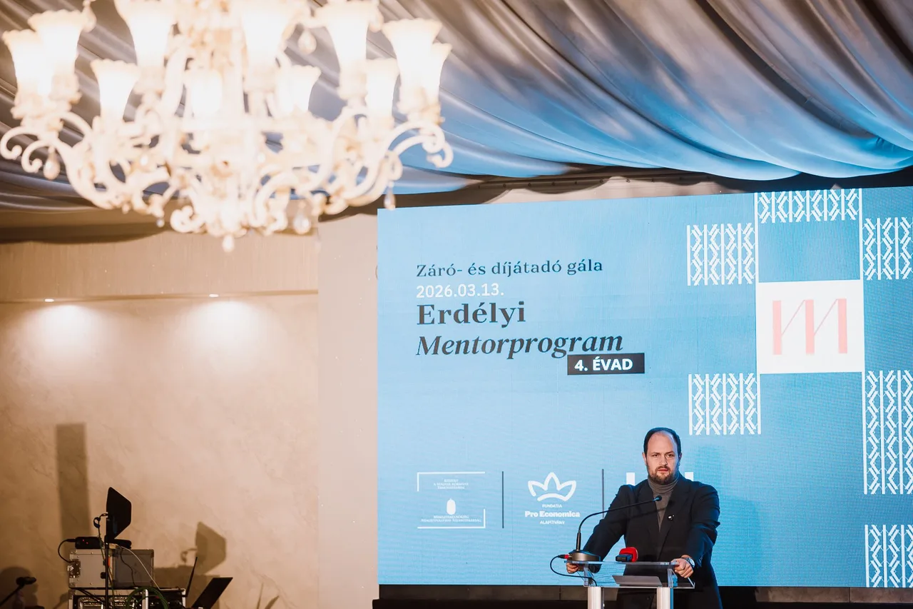 Öltönyt viselő férfi beszél egy pódiumon, mögötte nagy kék kivetítőn az „Erdélyi Mentorprogram – Záró- és díjátadó gála” felirat és a Pro Economica Alapítvány logója látható, a terem mennyezetén díszes kristálycsillár és kék drapéria díszíti a helyszínt.