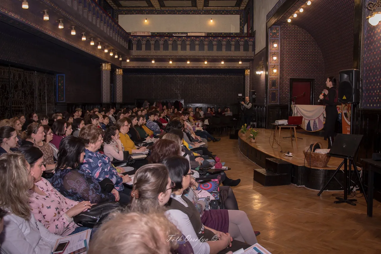 Előadóterem, ahol egy nő mikrofonnal a színpadon beszél a közönséghez; a nézőtéren nagyszámú, főként nőkből álló hallgatóság ül és figyel.