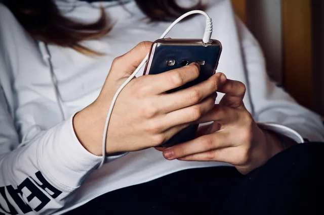 Közelkép egy fehér kapucnis pulóvert viselő személy kezeiről, aki egy okostelefont tart és kezel, miközben a telefon töltőkábellel van csatlakoztatva.