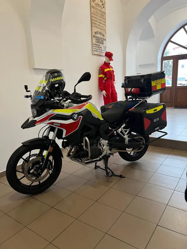 Sürgősségi mentőszolgálat (SMURD) feliratú BMW F800GS motorkerékpár egy előtérben, mögötte piros tűzoltó/paramedikus overallt viselő bábu és falra rögzített emléktábla látható.