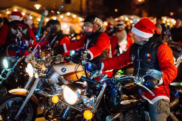 Motorosok mikulássapkás piros ruhában ülnek motorkerékpárjaikon éjszaka, karácsonyi fényfüzérekkel díszített téren, előtérben egy motoros románzászlós mellényén 'LIE' felirattal.