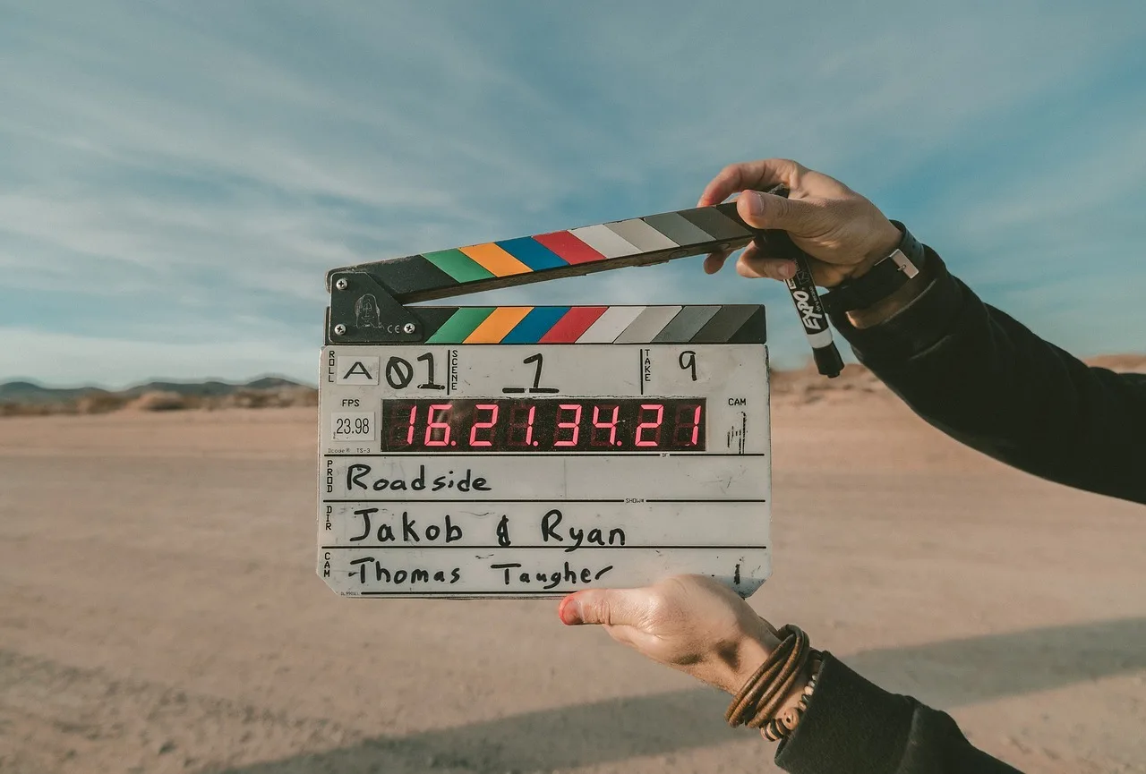 Egy személy filmes klapperdeszkát (csapódeszkát) tart a kezében, digitális időbélyeg-kijelzővel, kültéri, sivatagos-homokos táj és kék ég előtt. A táblán kézzel írt szöveg olvasható: Roadside, rendezők Jakob & Ryan, operatőr Thomas Taugher.