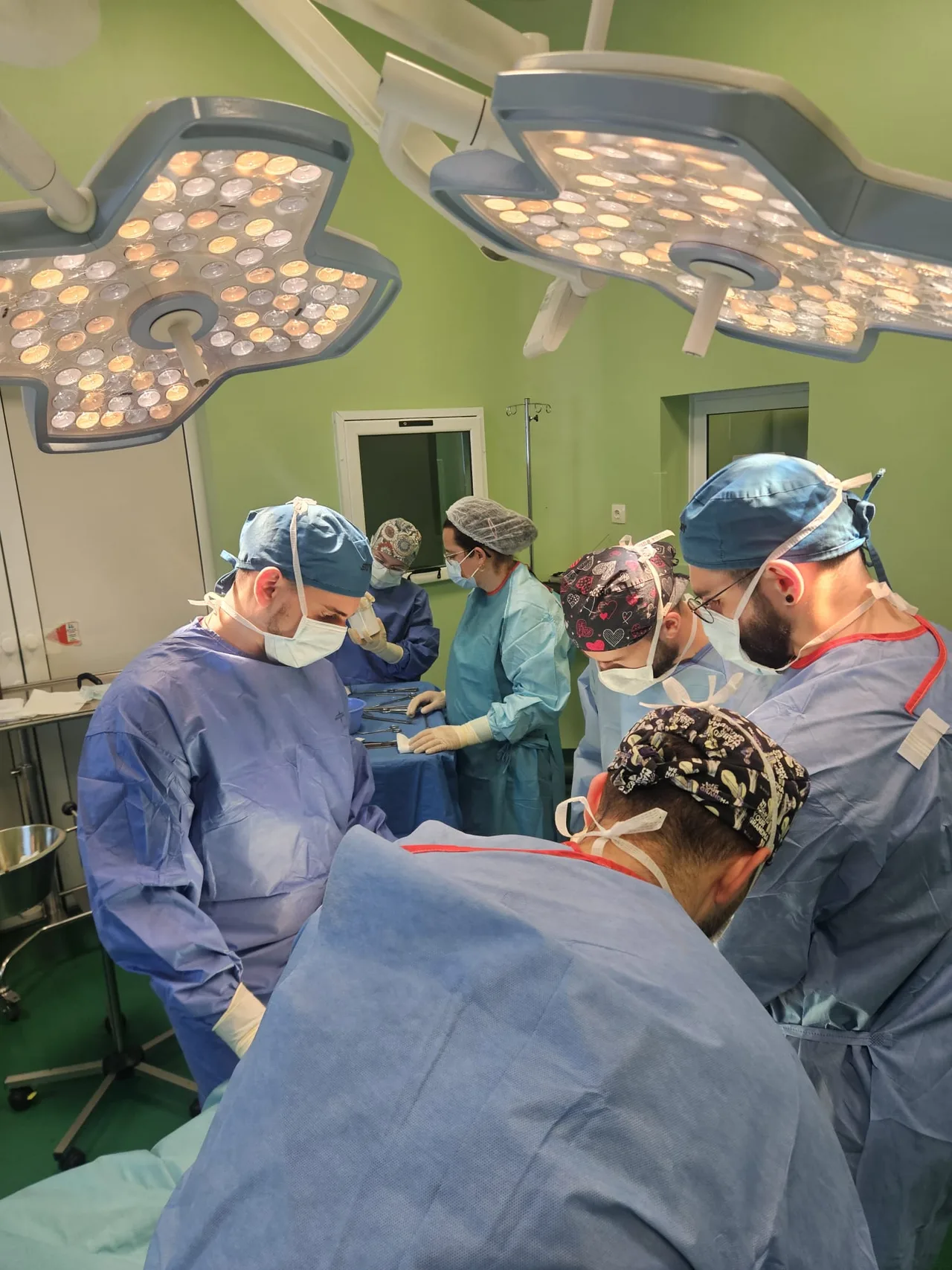 Műtőben több sebészeti maszkot és sapkát viselő orvos hajol egy operált beteg fölé, felettük két nagy műtőlámpa világít, a háttérben eszközasztalnál egy nővér dolgozik.
