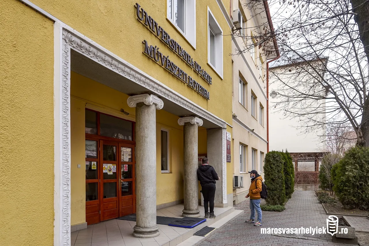 A Marosvásárhelyi Művészeti Egyetem sárga homlokzatú épületének bejárata, ión oszlopos portikusszal, felette a felirat román és magyar nyelven. A bejárat előtt egy férfi az ajtó felé tart, míg egy másik, sárga kabátos, sapkás és maszkos férfi az udvaron sétál.
