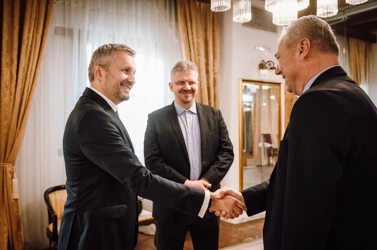 Két öltönyös férfi kézfogással köszönti egymást egy elegáns, csillárokkal és aranyszínű függönyökkel díszített teremben, mellettük egy harmadik öltönyös férfi figyeli a jelenetet.