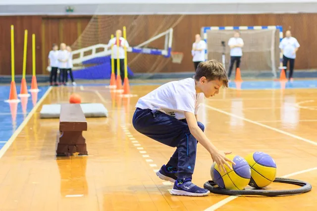 Sportcsarnokban egy fiú guggolva nyúl két sárga-kék labda felé, mögötte kúpokkal jelölt pálya és felnőttek állnak háttérben.
