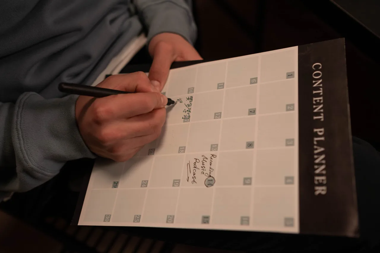 Közelkép egy kézről, amint filctollal ír egy „Content Planner” feliratú havi naptártervezőbe, amelyben számozott napokat tartalmazó rácsozat látható, néhány beírt jegyzettel.