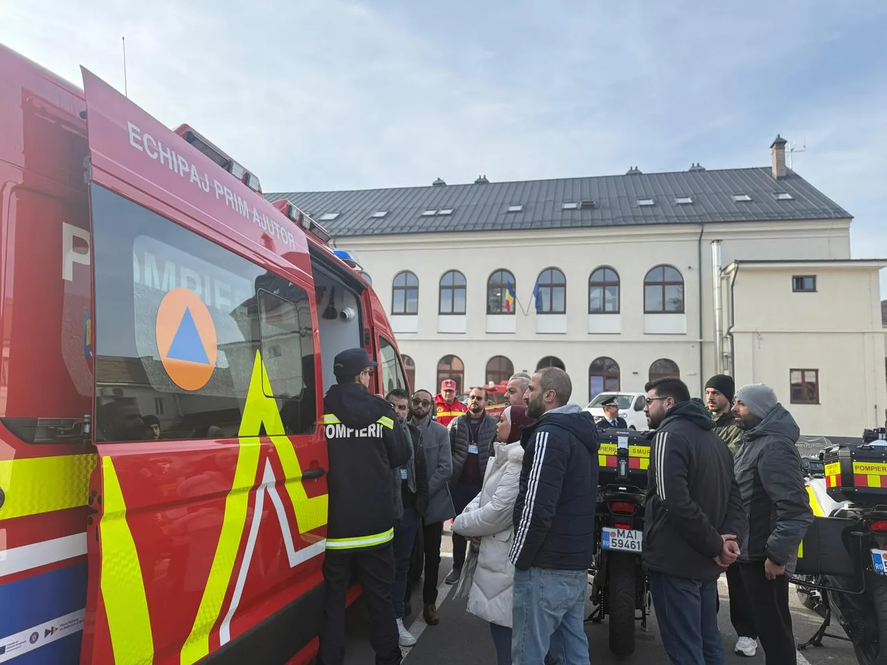 Piros tűzoltóautó előtt egyenruhás pomper magyaráz egy kisebb csoportnak, mögöttük egy régi, kétszintes világos épület és több tűzoltó motorkerékpár látható egy udvaron.