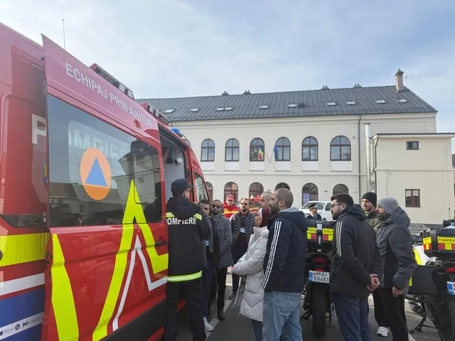 Piros tűzoltóautó előtt egyenruhás pomper magyaráz egy kisebb csoportnak, mögöttük egy régi, kétszintes világos épület és több tűzoltó motorkerékpár látható egy udvaron.