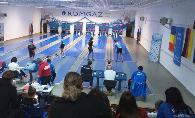 Fedett tekepálya, ahol több sportoló dobja a golyót a pályákon, a háttérben eredményjelző táblák és „ROMGAZ” felirat látható, előtérben nézők és edzők ülnek a padokon.