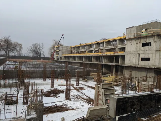 Építkezés télen: többszintes betonvázas épület alapozási munkálatai, előtérben hó borította talajon vasbeton armatúrák és építőanyagok, jobb oldalon a magasabb, több emeletes szerkezet, háttérben daru.
