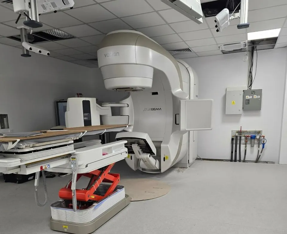 Belső orvosi szobában egy Varian TrueBeam típusú lineáris gyorsító (sugárterápiás) berendezés és egy hozzá kapcsolódó, hidraulikusan emelhető pácienságy látható. A fehér falú teremben felfüggesztett mennyezet, valamint falra szerelt elektromos és technikai szekrények, csövek figyelhetők meg.