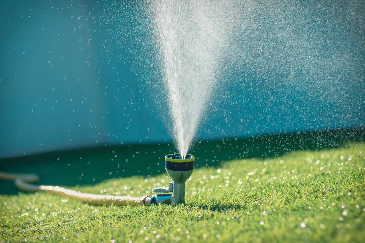 Egy locsolófejből erőteljes vízsugár permetezik zöld pázsitra napsütésben, a háttérben kék színű kerítés vagy fal látszik.