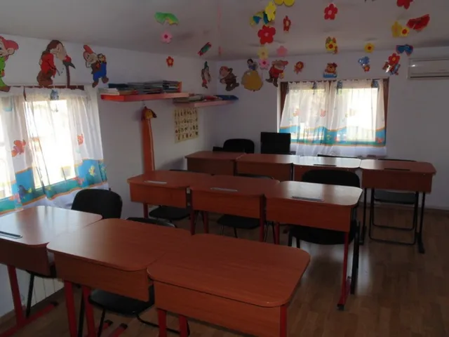 Üres tanterem barna faasztalokkal és fekete székekkel, a mennyezetről és falakról színes papírdíszek, virágok és mesefigurák lógnak, a falon polcokon könyvek és játékok láthatók.