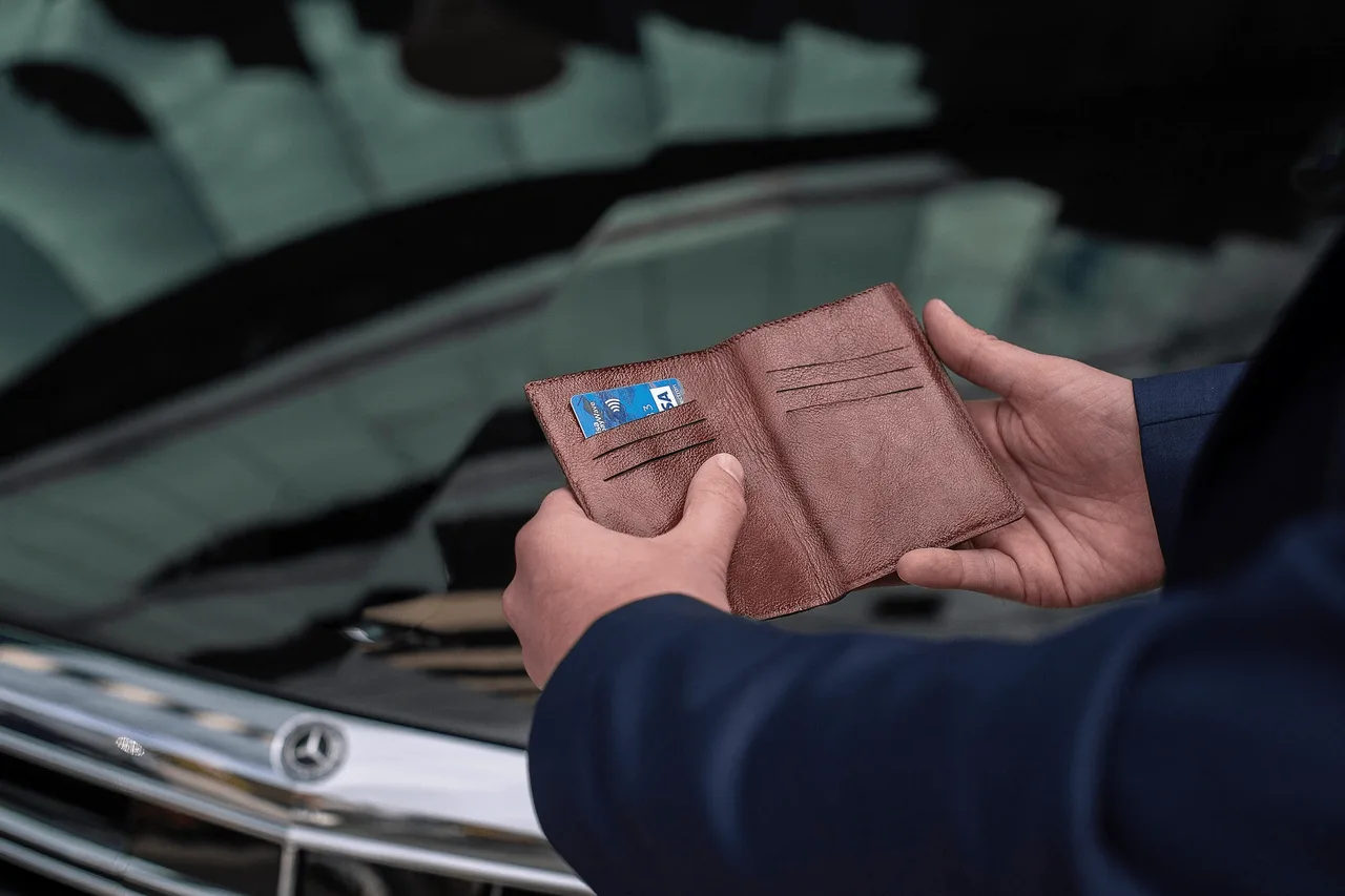 Egy férfi barna bőr pénztárcát tart a kezében, benne egy bankkártyával, egy autó motorháztetője előtt állva, a háttérben Mercedes embléma látszik.